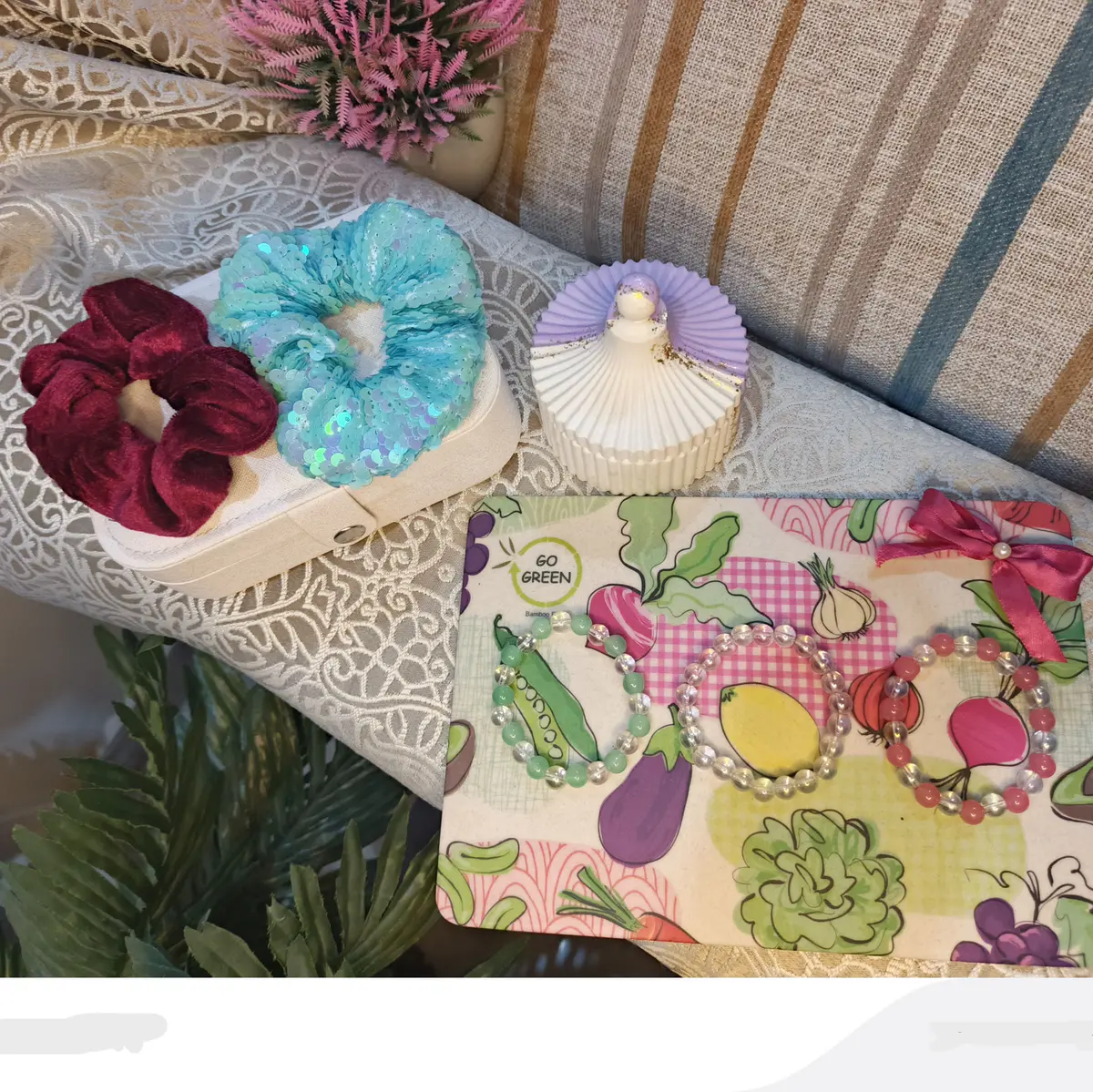 A soft, aesthetic flat lay featuring three handmade beaded bracelets and two scrunchies arranged on a lace surface. The bracelets have pastel pink, white, and green tones, while the scrunchies come in cozy velvet and shiny sequin textures. The setup includes a warm, decorative background for a delicate and feminine look.