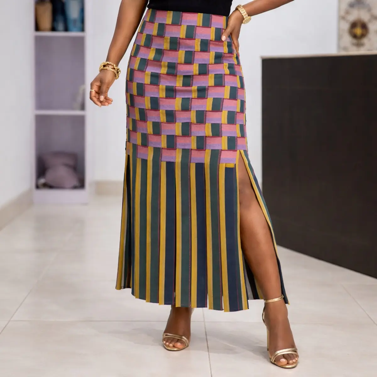Multicolor Ankara midi skirt with bold geometric patchwork design, high waist fit, front slit, and striped hem, styled with gold heels