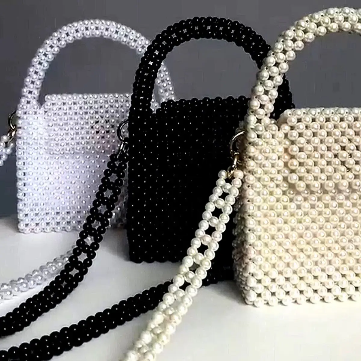 Three beaded mini handbags in white, black, and ivory with pearl-style beads, top handles, and crossbody straps displayed together