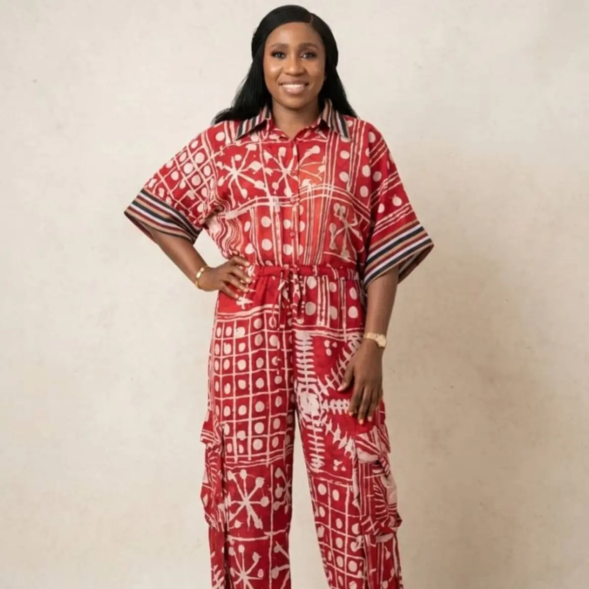 A closer front-facing view of the same handcrafted adire batik jumpsuit in deep crimson red and off-white, showcasing the intricate hand-dyed geometric and organic resist-print patterns across both the top and wide-leg cargo trousers. The striped collar and cuff trim are clearly visible against the bold red batik fabric. Color: Crimson Red and Off-White