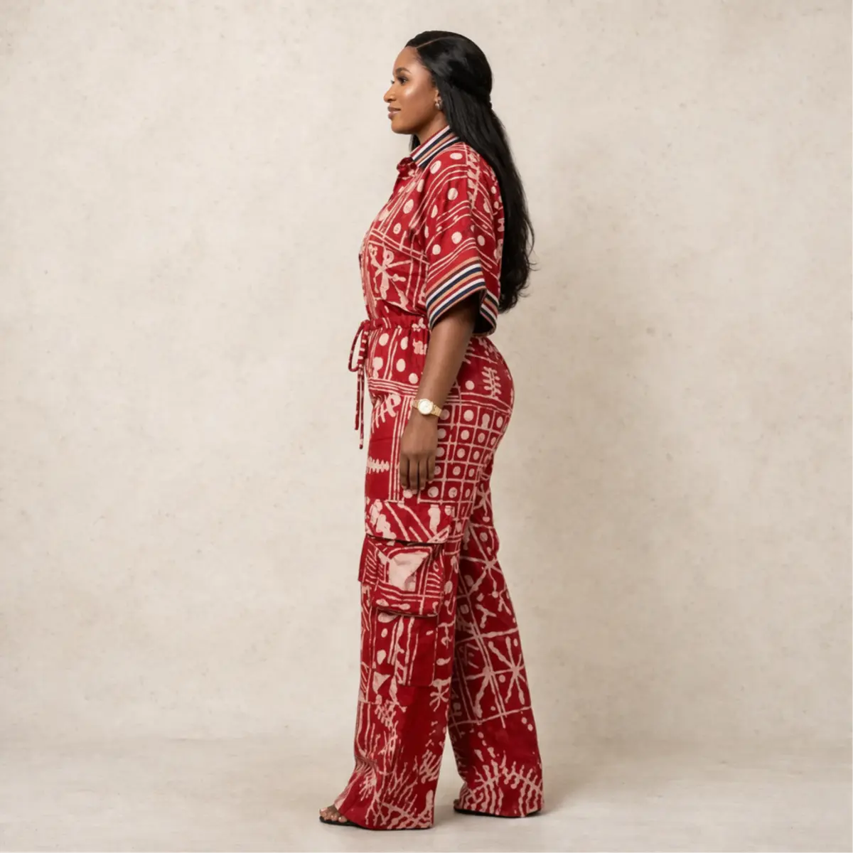 A side-profile view of the artisan-made adire batik wide-leg cargo jumpsuit in rich crimson red and off-white, highlighting the deep side cargo pocket on the trouser leg and the flowing wide-leg silhouette. The hand-dyed batik print with starburst and grid motifs is visible across the full length of the garment from this angle. Color: Crimson Red and Off-White
