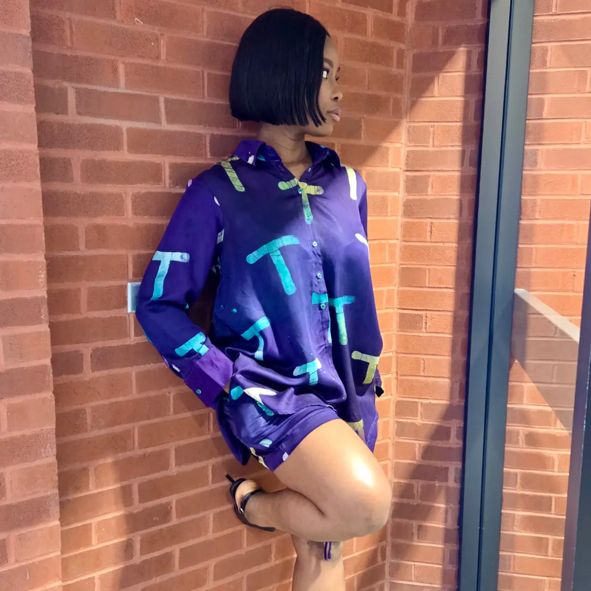 A woman poses against a red brick wall wearing a deep royal purple satin oversized button-down shirt with an allover hand-printed graphic motif in teal, lime green, and white, paired with matching purple shorts. The shirt features a classic collar and small buttons down the front. Color: Royal Purple