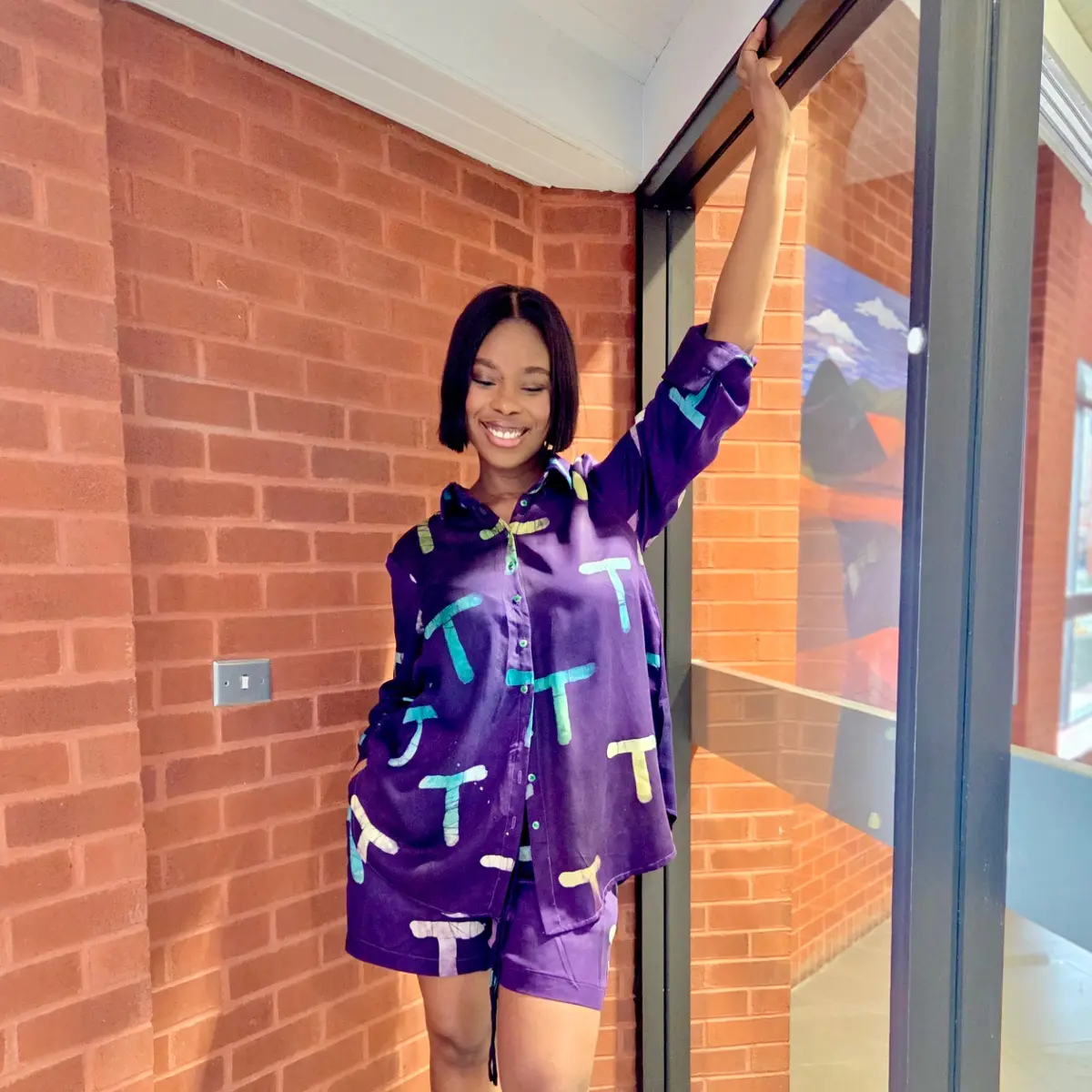 A smiling woman holds a glass door frame while wearing a vibrant purple satin two-piece co-ord set featuring a hand-printed allover graphic pattern in teal, lime, and cream tones on both the oversized shirt and the coordinating shorts. Color: Royal Purple