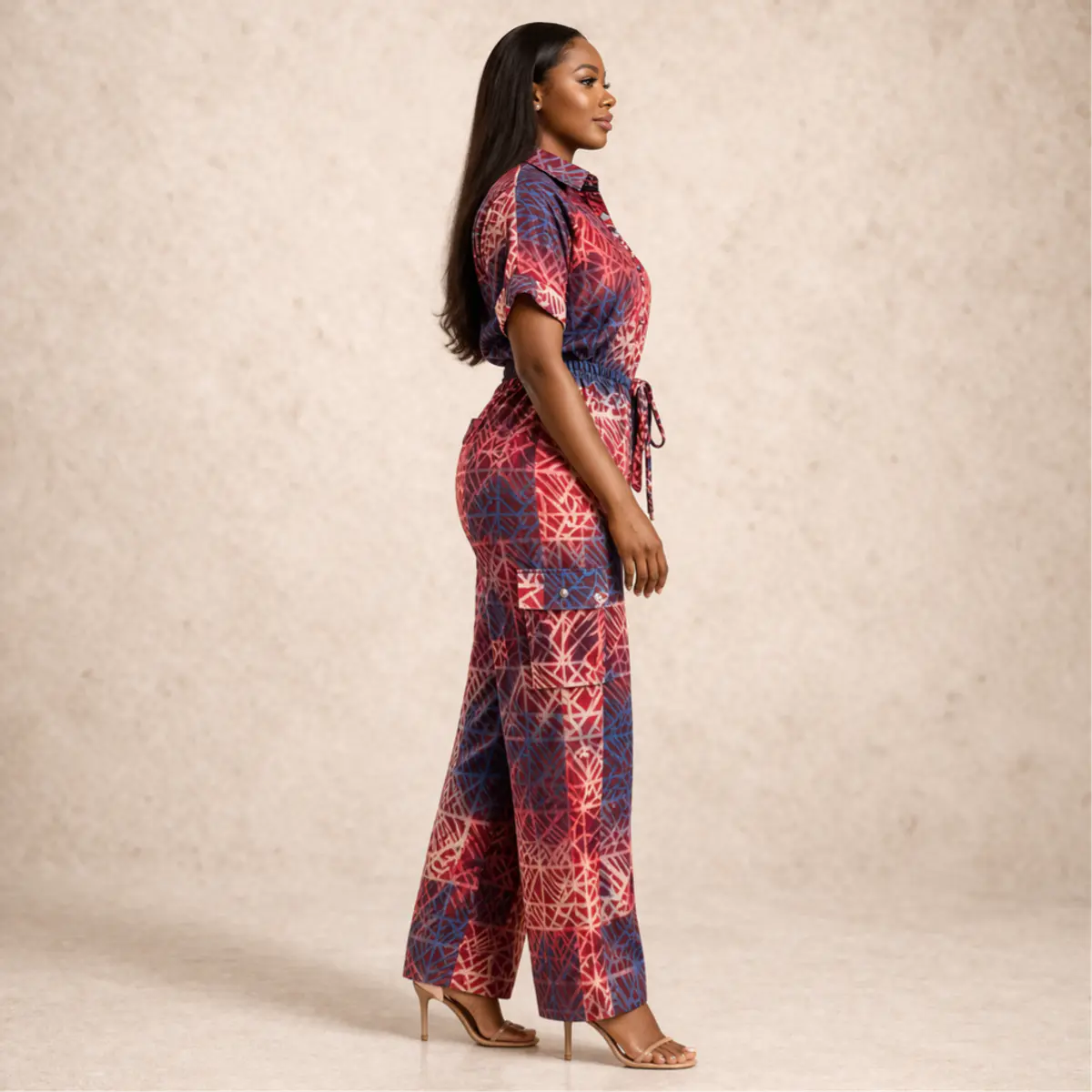 Side profile view of a woman wearing a handcrafted adire batik print wide-leg cargo jumpsuit in crimson red and deep navy blue. The side shot highlights the relaxed drape of the wide-leg trousers, the cargo pocket detail, and the beautifully balanced handcrafted geometric batik pattern flowing across the full garment. Color: Crimson Red and Navy Blue