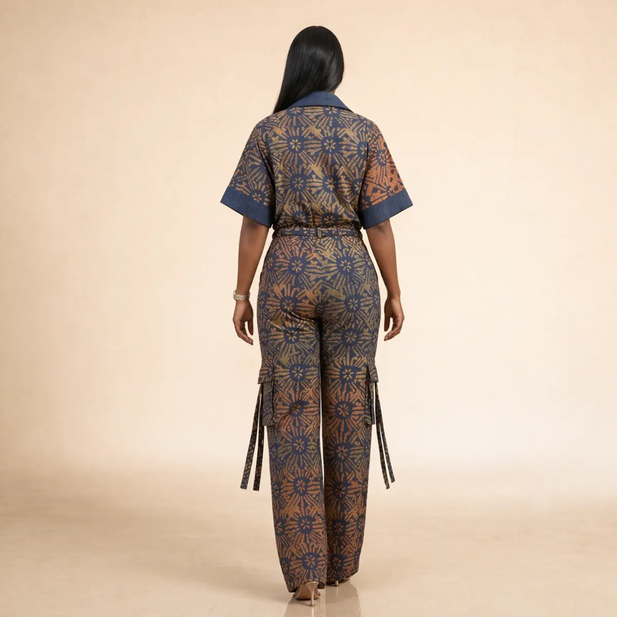 Rear view of a woman wearing the full handcrafted Adire batik co-ord set, highlighting the back silhouette of the boxy shirt with denim-trim cuffs, the drawstring waistband, and the wide-leg cargo pants featuring dual utility pockets with decorative hanging fabric tie straps in the matching navy blue, burnt gold, and terracotta batik print. Color: Navy Blue, Burnt Gold, Terracotta