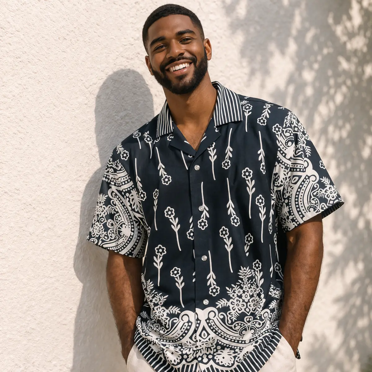 Close-up front view of a smiling man wearing a handcrafted short-sleeve button-up shirt in deep navy blue and white. The shirt features a bold artisan-made print combining paisley medallion borders at the sleeves and hem with delicate hand-embossed-style floral and botanical stem motifs across the body, and a contrast stripe collar. Color: Navy Blue and White