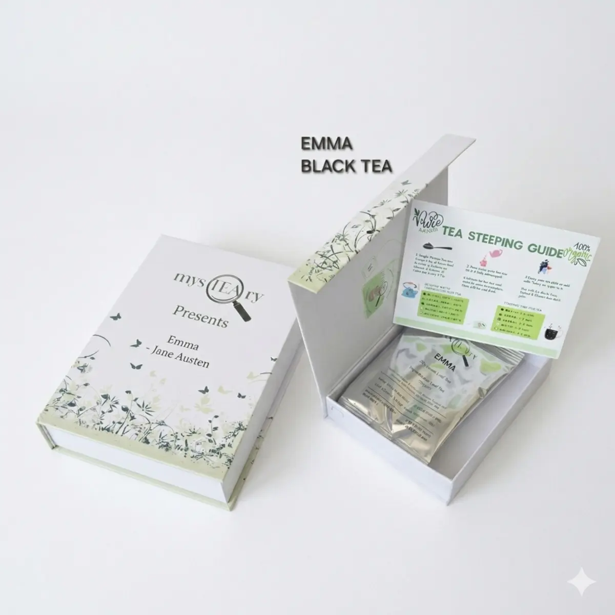 A flat-lay and angled product shot of the Emma Black Tea gift box, styled to resemble a classic hardcover novel with soft sage green and white botanical print packaging. The open box reveals a sealed foil pouch of loose-leaf tea and a Tea Steeping Guide card, presented as a one-of-a-kind unique handmade gift set. Color: Sage Green and White
