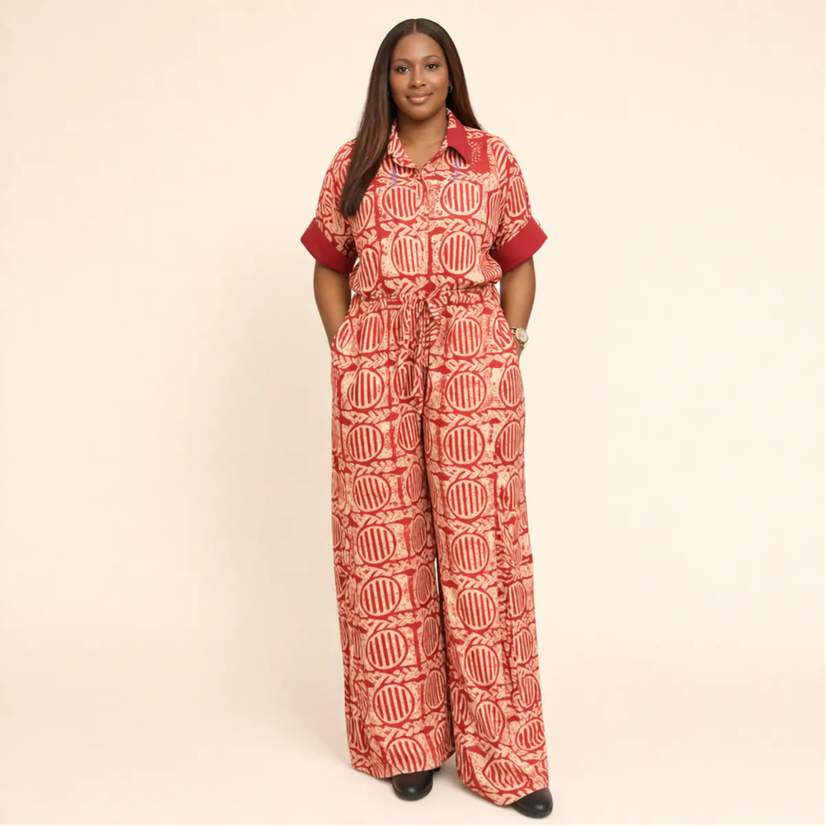 Full-length front view of a woman wearing an artisan-made African batik wide-leg jumpsuit in a striking red and cream colorway, showcasing the floor-grazing palazzo trouser silhouette, drawstring waist, and all-over circular batik print. The handcrafted garment drapes beautifully from shoulder to floor. Color: Red and Cream