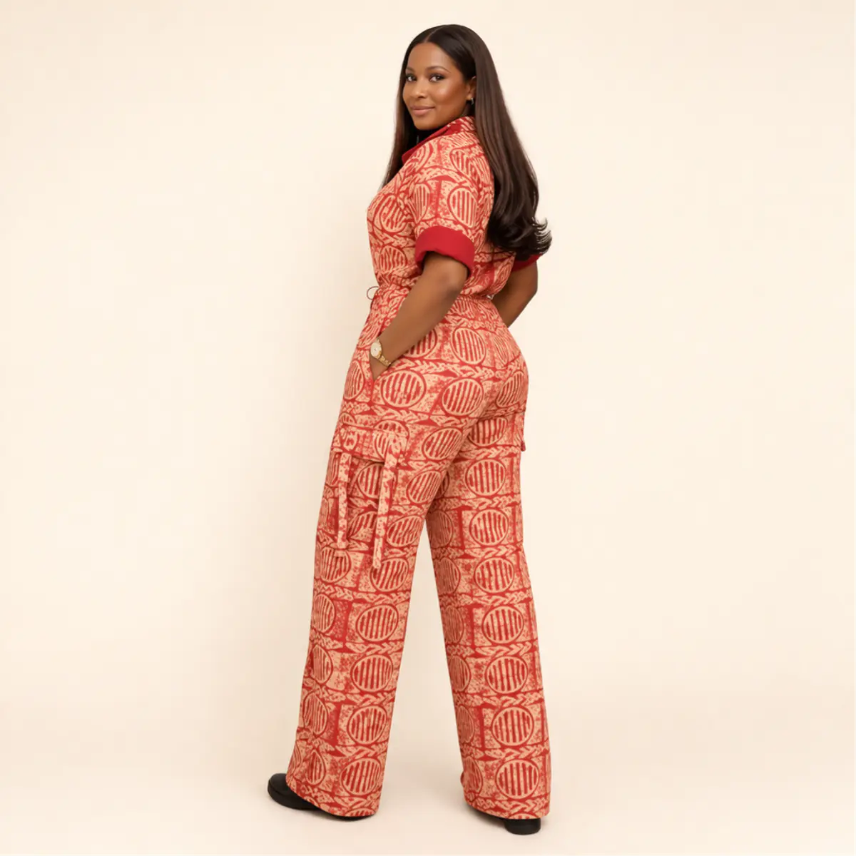 Rear three-quarter view of a woman wearing a handcrafted African batik print wide-leg jumpsuit in red and cream, highlighting the clean back silhouette, self-tie drawstring waist detail, and the bold all-over geometric batik pattern on the flowing wide-leg trousers. Color: Red and Cream