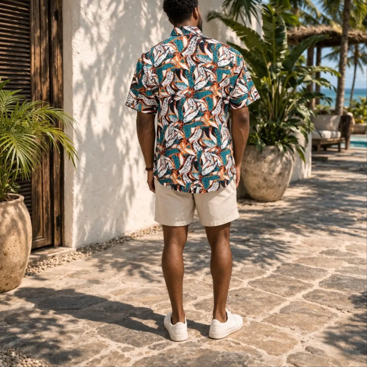 A rear view of a man walking along a tropical villa pathway, displaying the full back of the handcrafted African print camp shirt with its vibrant all-over teal, burnt orange, white, and black abstract motif, paired with cream shorts and white sneakers. The seamless artisan-made print wraps boldly across the back, confirming the small-batch quality and one-of-a-kind character of this piece. Color: Teal, Burnt Orange, White, and Black