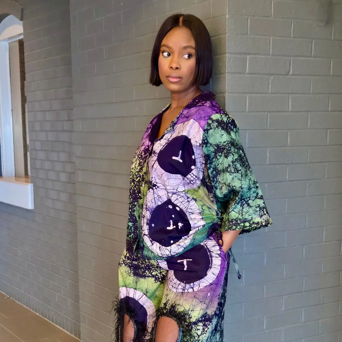Side profile view of a woman wearing a handcrafted African batik two-piece set featuring a kimono-style oversized top and wide-leg trousers. The fabric displays a striking wax-resist batik pattern in deep purple, forest green, black, and pale lavender with bold circular motifs and crackle detailing. Feather trim is visible along the lower hem of the trousers. Color: Purple, Green, Black, Lavender