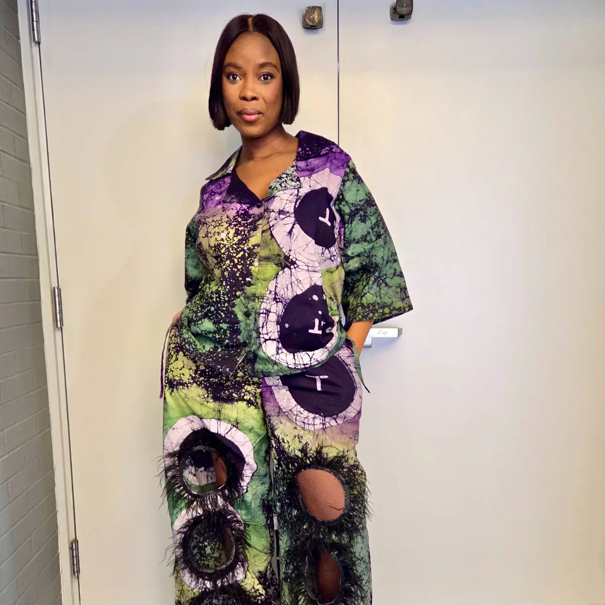 Front-facing full-length view of a woman wearing the handcrafted African batik co-ord set, showcasing the open-collar top and wide-leg pants adorned with large circular batik medallions in black surrounded by green, purple, and pale lavender crackle-dyed wax print fabric. Black feather trim details are visible at the trouser hem and circular cutout accents. Color: Purple, Green, Black, Lavender