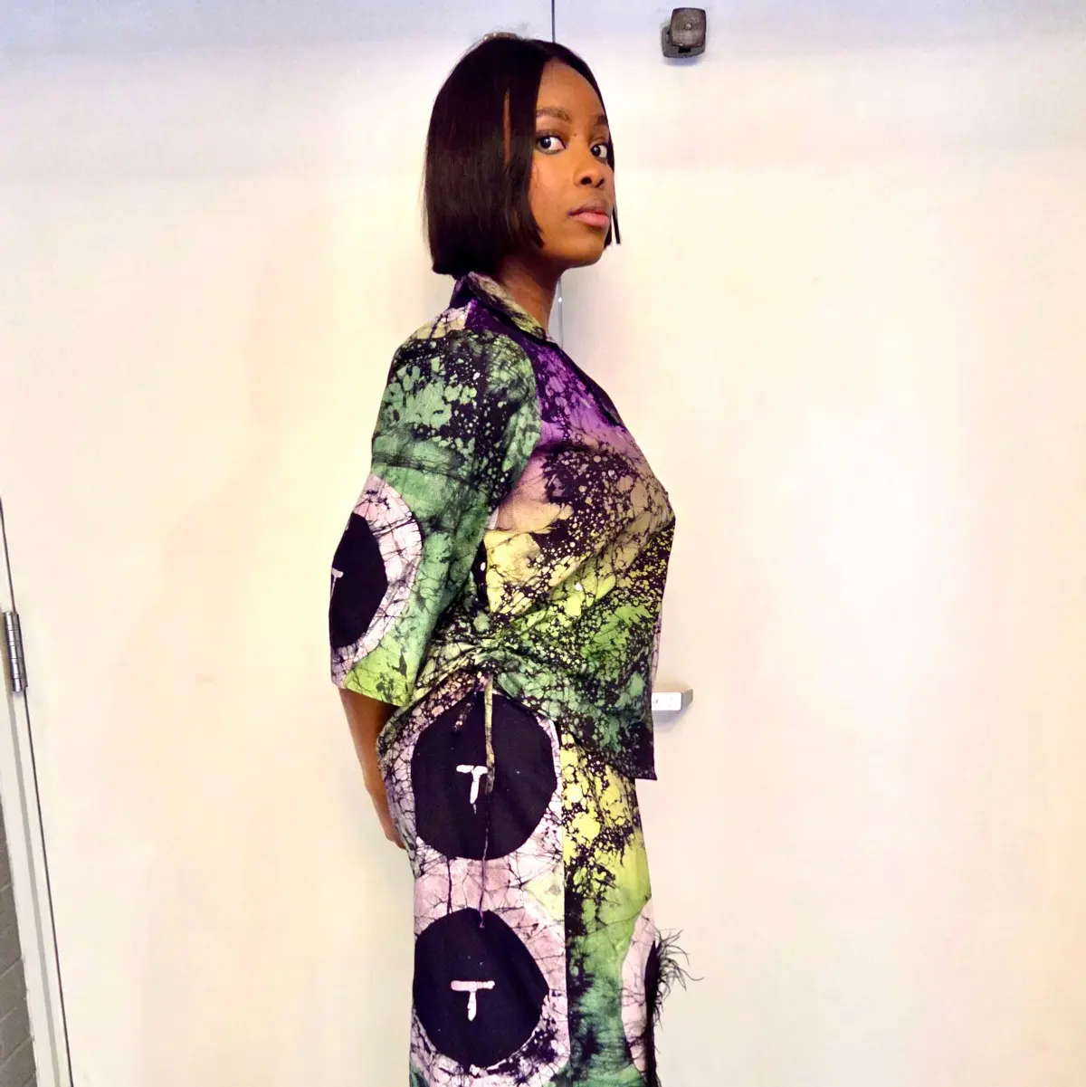 Side profile close-up of the handcrafted African batik two-piece set highlighting the fluid drape of the wide-leg trousers and kimono top. The wax-resist batik print features large black circular medallions against a vivid background of lime green, deep purple, and soft lavender crackle patterns, with feather trim peeking at the trouser hem. Color: Purple, Green, Black, Lavender