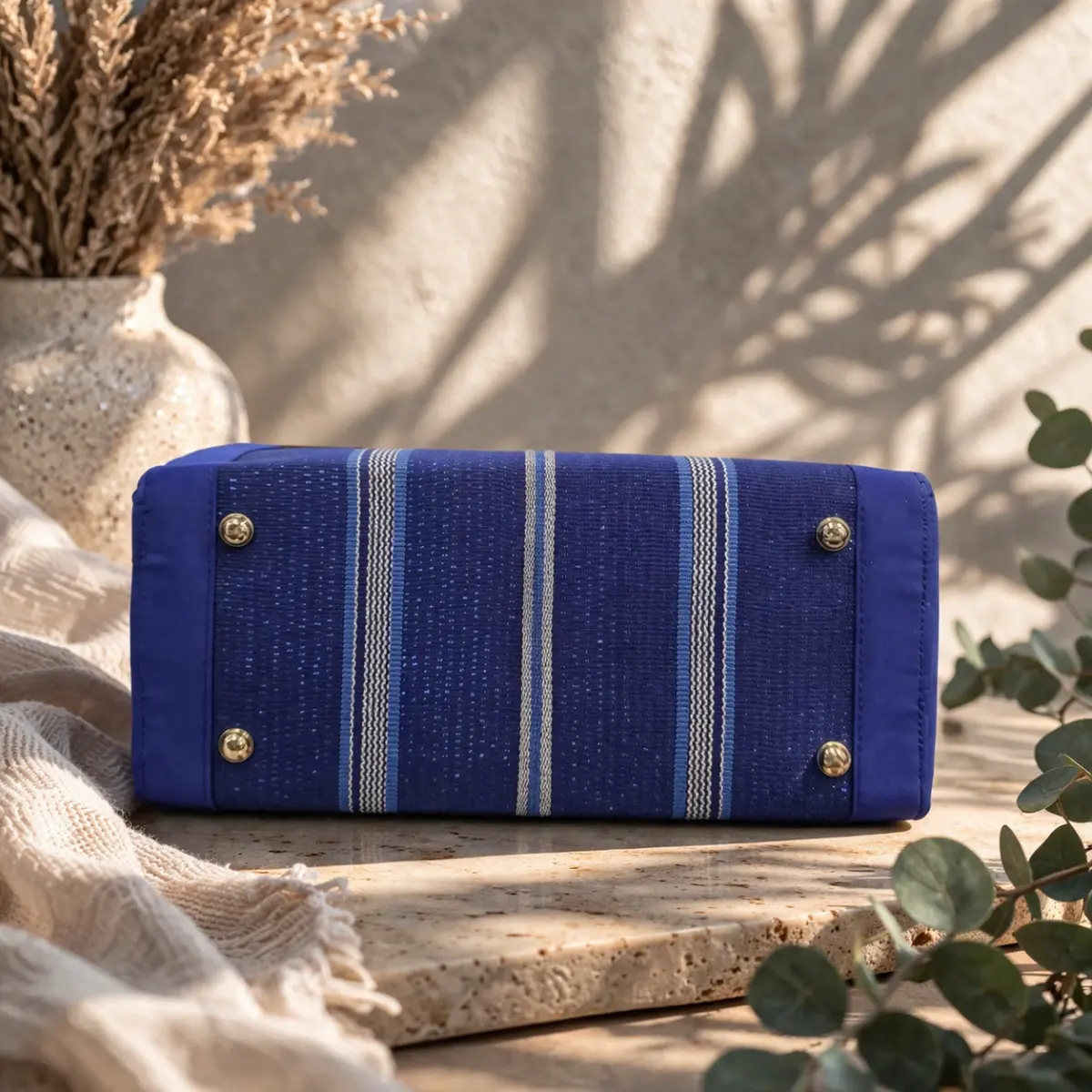 Bottom view of a handwoven Aso Oke tote bag lying flat on a travertine surface, showcasing its richly textured African fabric with bold vertical stripes in navy, sky blue, and cream. The bag features meticulous artisan stitching along the edges and four protective gold-tone metal feet, highlighting the skilled craftsmanship behind this handmade piece. Styled alongside eucalyptus sprigs and dried pampas grass for an earthy, elegant aesthetic. Color: Navy Blue, Sky Blue, Cream, Gold