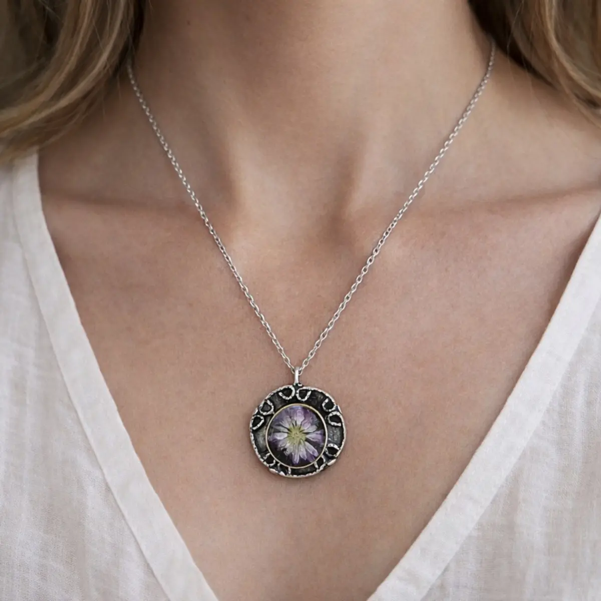 A woman wearing a handcrafted round pendant necklace on a delicate silver-toned stainless steel chain, resting at the neckline of a white V-neck top. The artisan pendant features a real dried purple chrysanthemum flower preserved in clear resin, set within an ornate silver-plated brass bezel with a textured, lace-like decorative border. The botanical centerpiece showcases the flower's intricate petals with a pale yellow core, highlighting skilled handmade craftsmanship. Color: Silver, Purple, White, Yellow