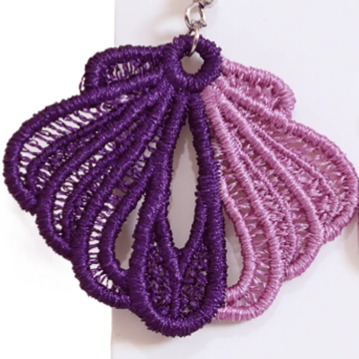 Extreme close-up detail shot of a single handmade lace butterfly wing earring propped against a white surface, highlighting the delicate hand-embroidered thread texture, layered fan-shaped lace structure, and the two-tone contrast between deep violet and pale mauve pink. Color: Purple and Mauve Pink