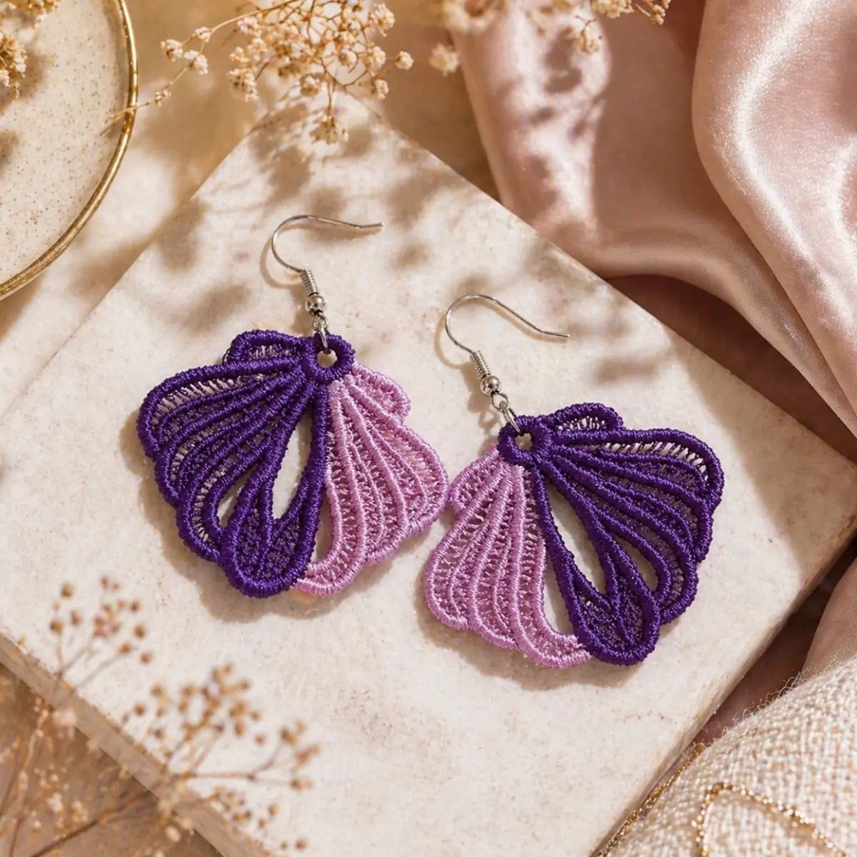 A pair of handcrafted lace earrings featuring an intricate fan or seashell-shaped design with delicate open threadwork. Each earring showcases a two-tone artisan embroidery style, combining deep violet and soft blush pink threads in a radiating ribbed pattern, finished with silver-tone fishhook clasps. The lightweight textile construction highlights skilled craftsmanship typical of handmade textile jewelry. Color: Purple and Pink