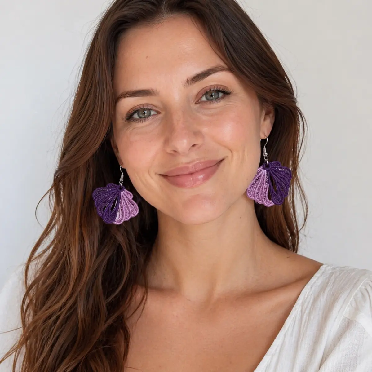 A smiling woman models a pair of handcrafted drop earrings featuring an elegant seashell or fan-shaped design crafted from delicately layered soft threads. The artisan earrings showcase intricate ribbed textile work in a two-tone color combination, suspended from silver-tone fishhook wires and measuring approximately 3.5 cm long by 4 cm wide. The careful thread-wrapping technique highlights skilled handmade craftsmanship, making these a chic statement accessory. Color: Purple and Lilac Pink