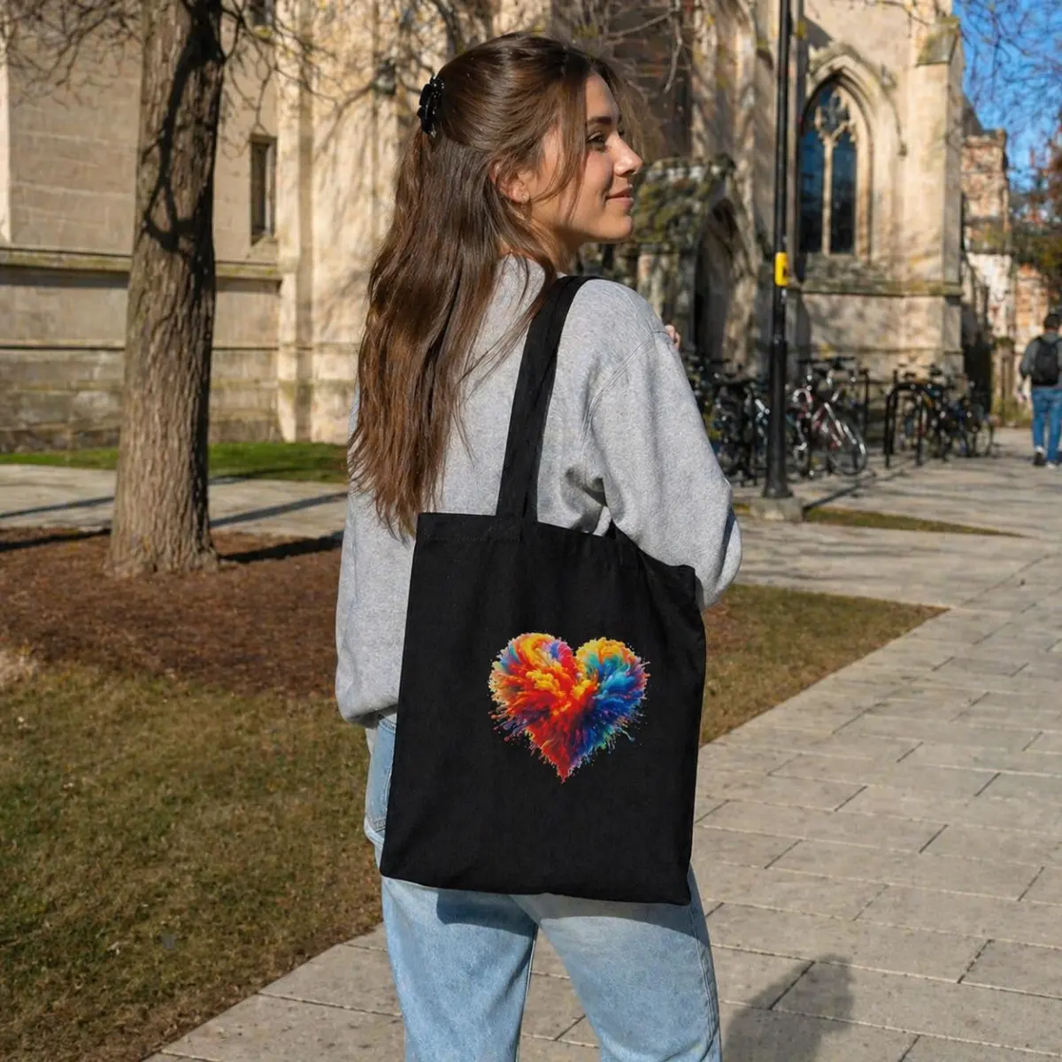 A young woman carrying a black canvas tote bag over her shoulder outdoors on a stone-paved university walkway, wearing a grey sweatshirt and jeans. The artisan-designed heart motif in fiery reds, oranges, yellows, and cool blues pops brilliantly against the deep black background of the bag. Color: Black with Multicolor Heart Print