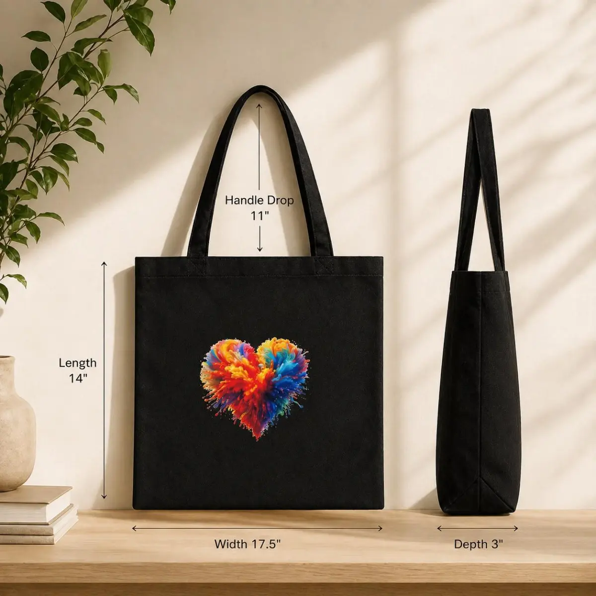 A flat lay and side-profile product shot of the black canvas tote bag on a wooden surface, showing exact dimensions — 17.5 inches wide, 14 inches long, 3 inches deep, and 11-inch handle drop — with the vivid multicolor heart graphic centered on the front panel. Color: Black with Multicolor Heart Print