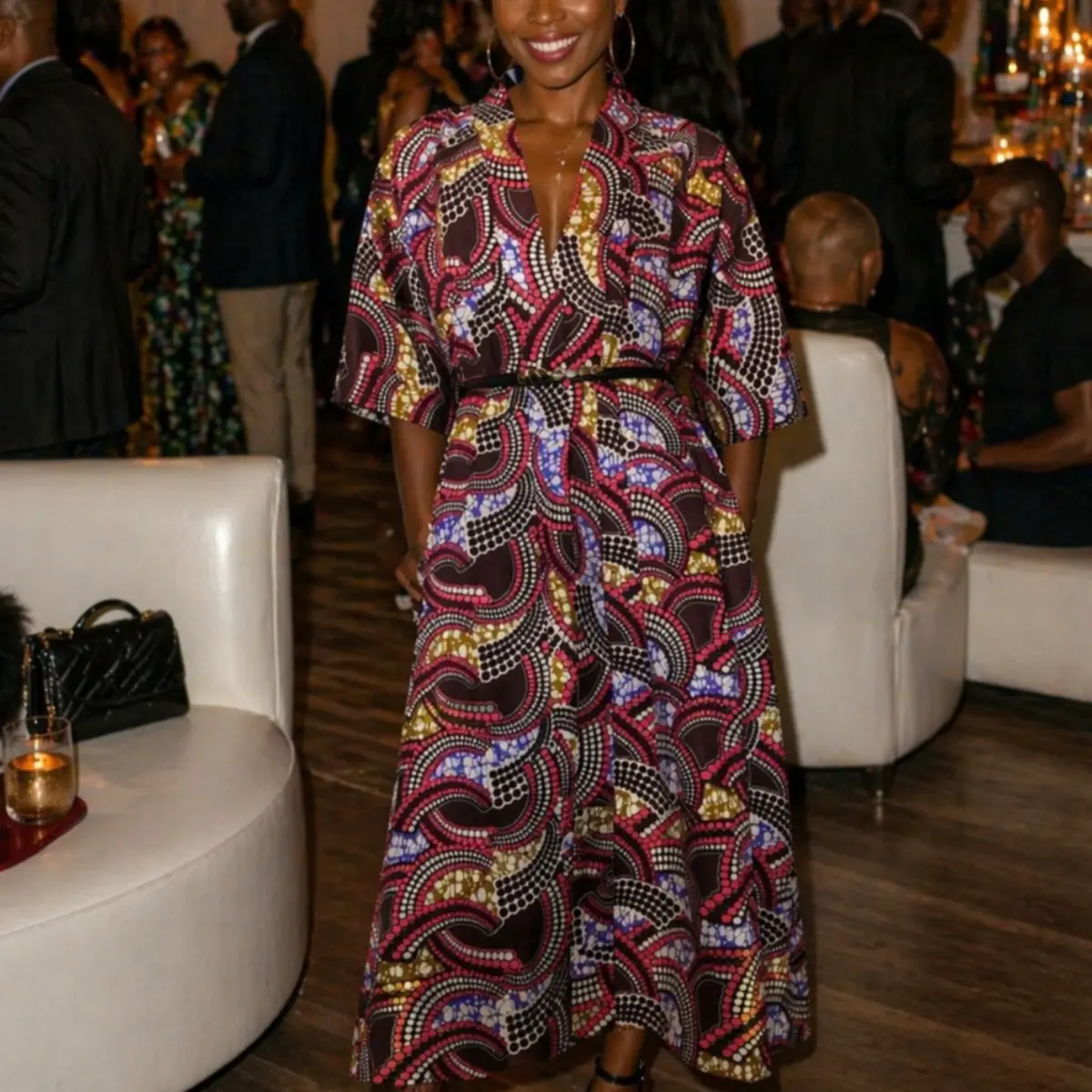 A woman wearing a handcrafted Ankara wax print midi wrap dress with a deep V-neckline, three-quarter sleeves, and a cinched waist, styled for an evening event. The fabric features a bold swirling pattern in deep burgundy, cobalt blue, olive gold, pink, and white on a dark brown ground. Color: Burgundy Multi