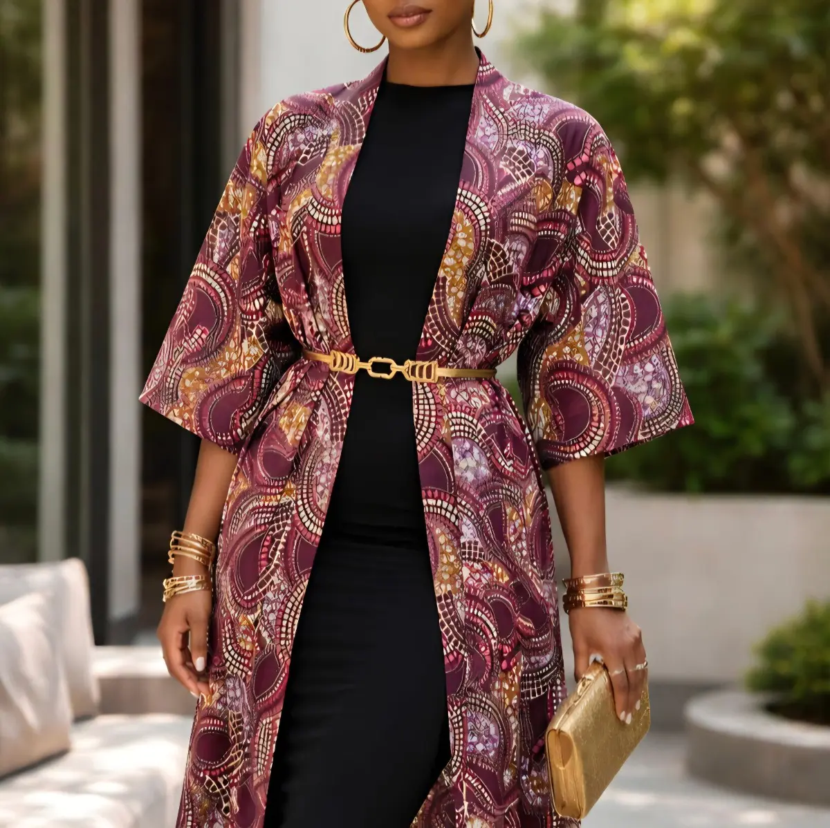 Close-up of a woman wearing a handcrafted Ankara wax print longline kimono duster layered over a fitted black dress, cinched with a gold belt and accessorized with gold bangles and a gold clutch. The kimono features a rich plum and mauve swirling paisley motif with gold, pink, and ivory accents. Color: Plum Mauve Gold