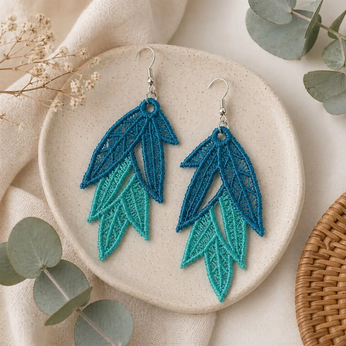 A pair of handcrafted lace earrings displayed on a speckled ceramic dish, featuring an intricate leaf-cluster design with delicate open-weave embroidery. The earrings showcase a two-tone gradient transitioning from deep ocean blue at the top to soft turquoise at the pointed tips, measuring 5.5 cm × 2.5 cm and finished with silver-tone fish-hook findings. Styled alongside dried baby's breath and eucalyptus sprigs, these artisan textile earrings reflect skilled needlework inspired by sea and sky. Color: Deep Blue, Turquoise